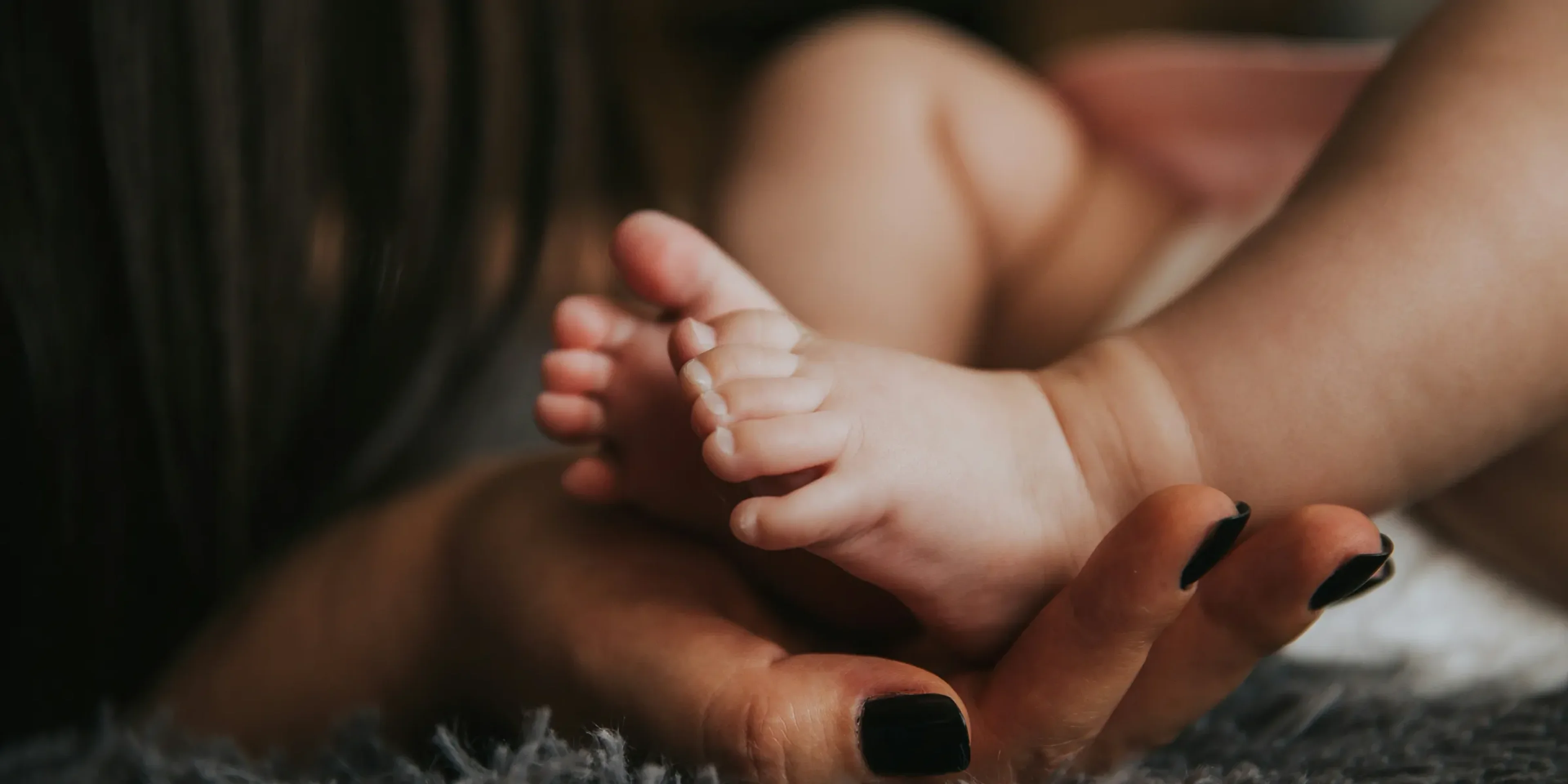 Infant's feet being held by a woman's hand with painted and manicured hands resting on a gray blanket