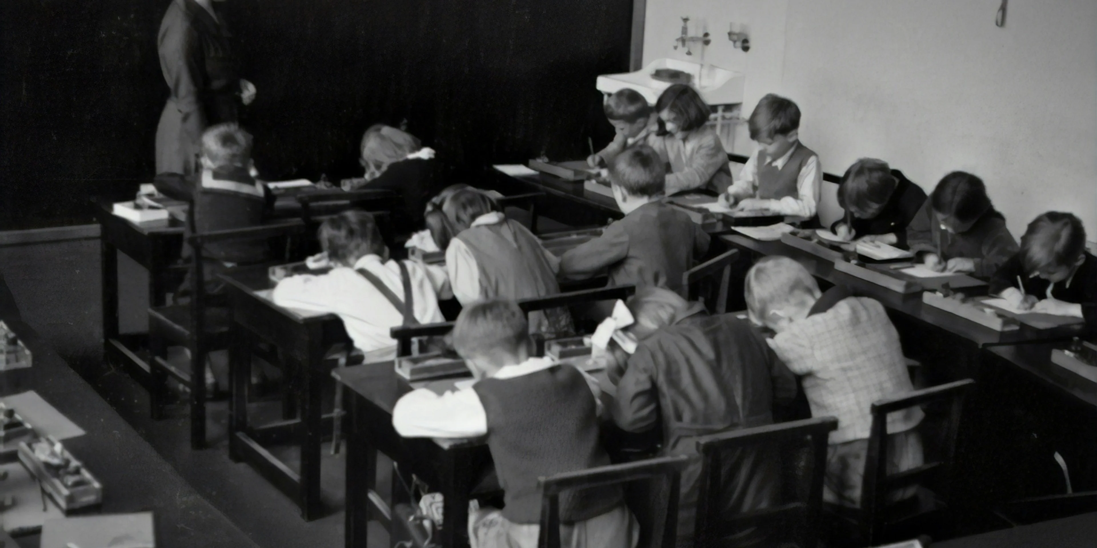 grayscale photography of teacher standing near chalkboard and children sitting on chairs.